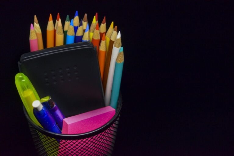 A collection of colored pencils, pens, and an eraser in a black mesh holder.