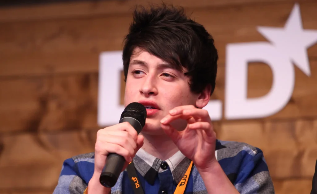 Young man speaking into a microphone during a panel discussion, wearing a checkered shirt and gesturing with his hands.