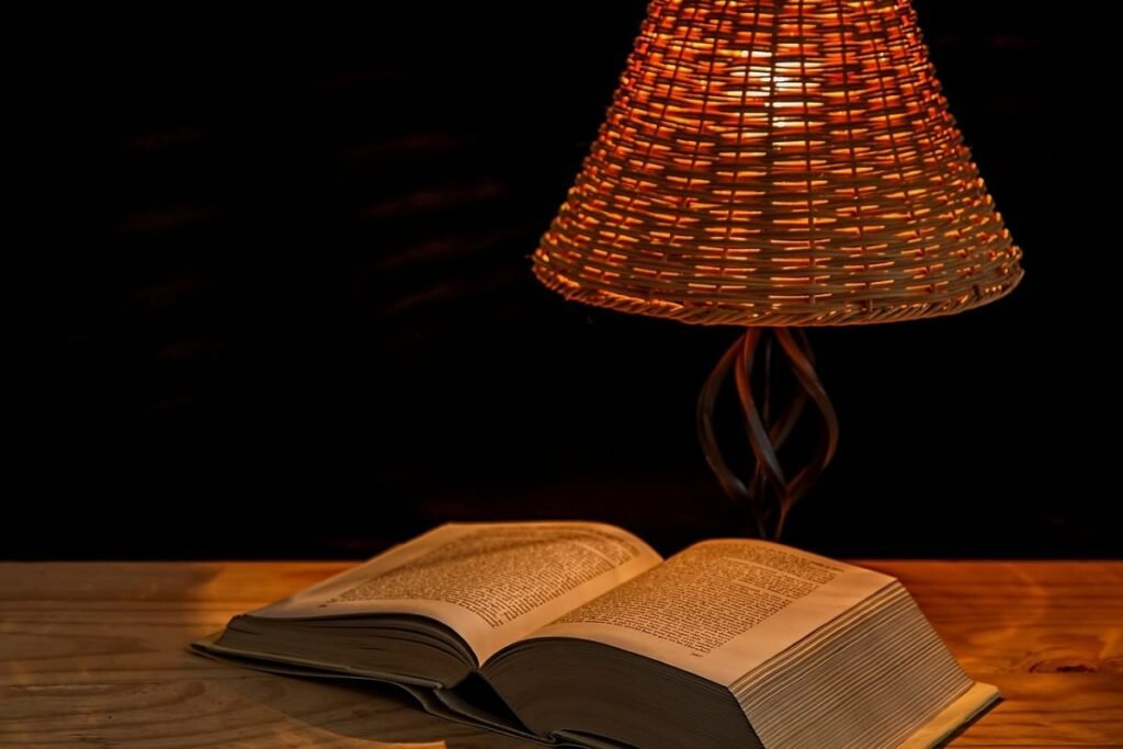 An open book resting on a wooden surface beside a woven lamp emitting warm light.
