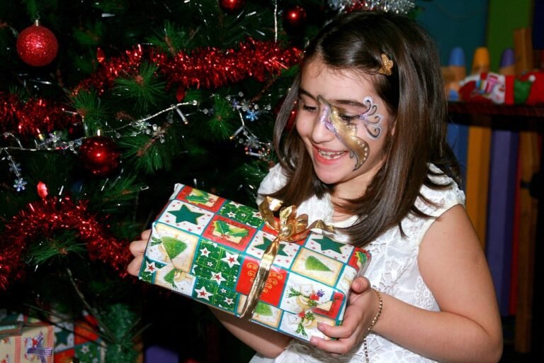 Child with face paint holding a wrapped gift in front of a decorated Christmas tree.