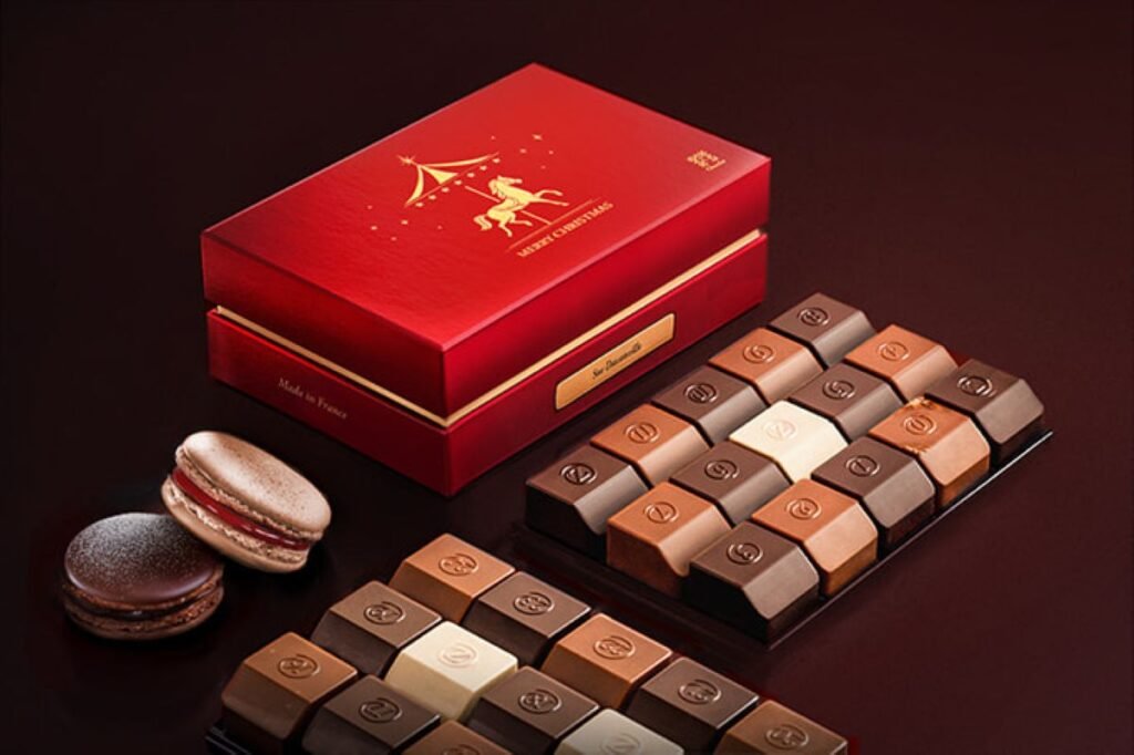 Red luxury chocolate gift box with assorted chocolates and macarons arranged neatly on a dark surface.