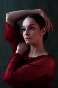 Portrait of a young woman with a thoughtful expression, wearing a red sheer blouse and posing with her arms raised.