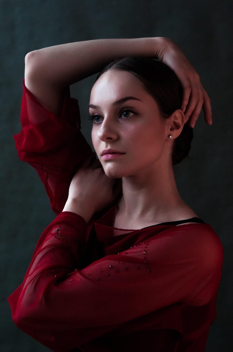 Portrait of a young woman with a thoughtful expression, wearing a red sheer blouse and posing with her arms raised.