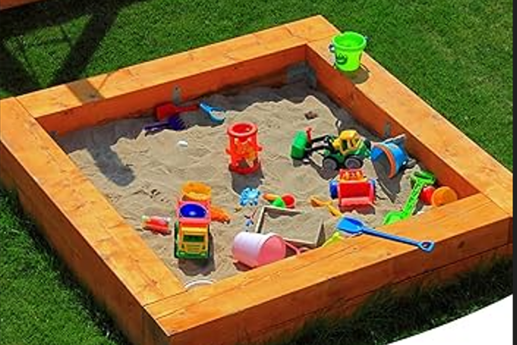 Sandbox with a wooden frame filled with sand and colorful plastic toys in a sunny backyard, including buckets, shovels, and toy vehicles.